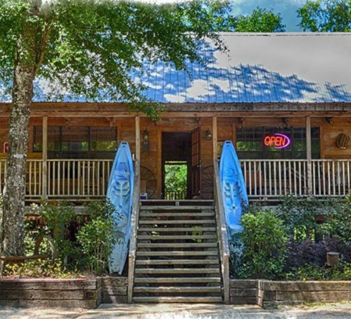 A rustic wooden cabin with a blue roof, featuring an "Open" sign and two kayaks by the entrance surrounded by greenery.