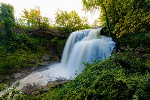 A serene waterfall cascades over rocks, surrounded by lush green trees and foliage in a tranquil natural setting.