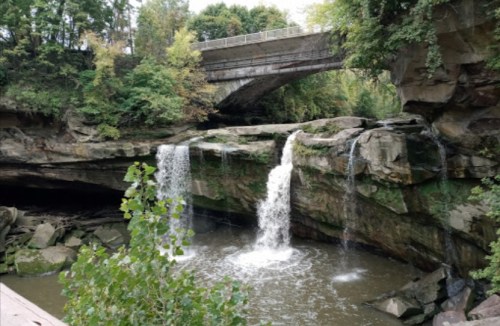 A serene waterfall cascades over rocky cliffs beneath a bridge, surrounded by lush greenery.