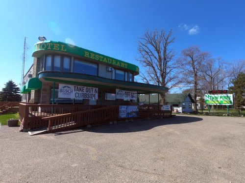 A motel and restaurant with a green awning, featuring signs for takeout and curbside service, set against a clear blue sky.