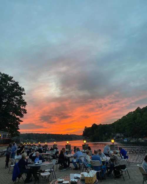 Outdoor dining scene by a lake at sunset, with people seated at tables and colorful clouds in the sky.
