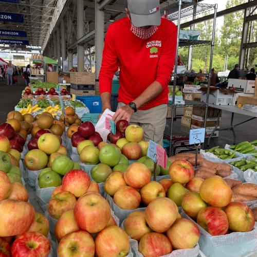 A vendor arranges apples and other produce at a bustling farmers market. Bright colors and fresh fruits are on display.