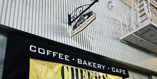 Sign for a café and bakery featuring "Coffee, Bakery, Café" and "Cheers" on a storefront with a decorative hanging sign.