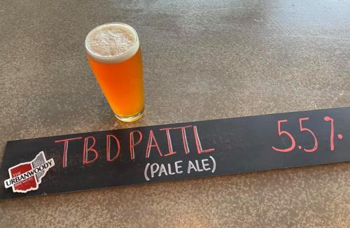 A glass of pale ale sits next to a chalkboard sign reading "TBD PAITL" and the price of $5.57.