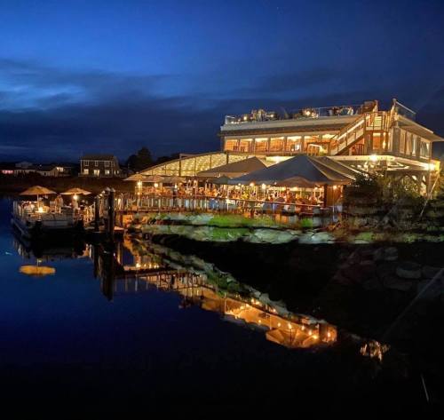 A waterfront restaurant illuminated at night, reflecting in calm water, with outdoor seating and a cozy atmosphere.