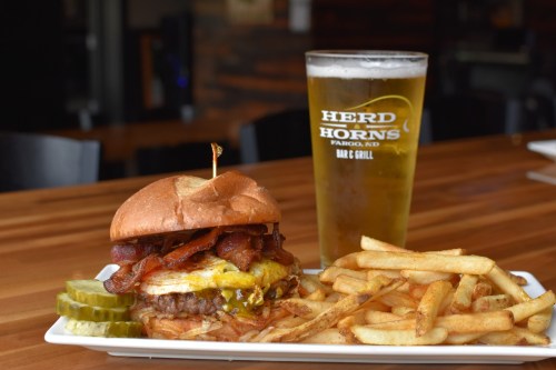 A juicy burger with bacon and cheese, served with fries and pickles, alongside a pint of beer in a branded glass.