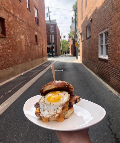 A hand holds a plate with a sandwich featuring a fried egg, bacon, and cheese, set against a brick alleyway.