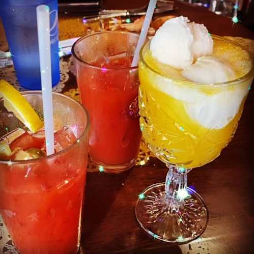Three colorful drinks on a table: a red cocktail with a lemon slice, a red drink, and a yellow drink topped with ice cream.