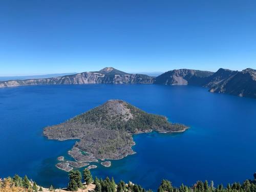 A panoramic view of Crater Lake, featuring a blue lake, an island, and surrounding cliffs under a clear blue sky.
