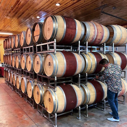 A person inspects wine barrels stacked in a winery, with wooden ceilings and warm lighting in the background.