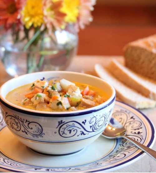A bowl of soup with vegetables and chicken, served on a decorative plate, with bread slices and flowers in the background.