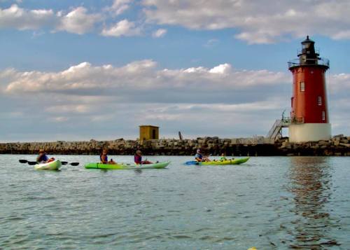 Four kayakers paddle in calm waters near a red lighthouse under a cloudy sky.