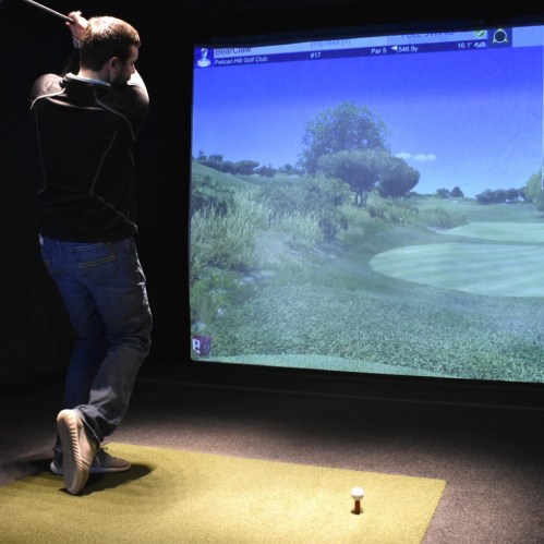 A golfer swings a club in a simulator, with a virtual golf course displayed on a large screen.