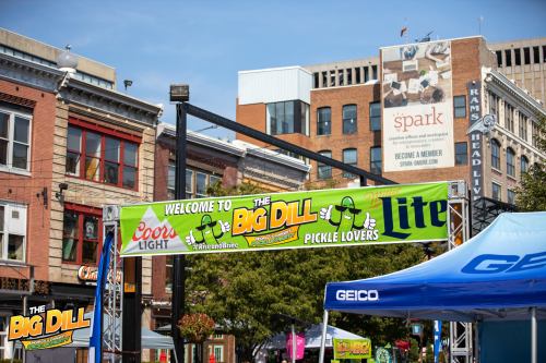 A colorful banner reads "Welcome to The Big Dill" at a festival, with buildings and tents in the background.