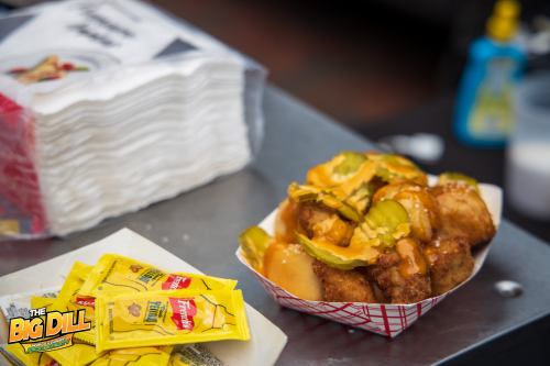 A serving of fried food topped with pickles and cheese sauce, alongside packets of mustard on a metal surface.