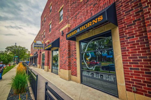 Exterior view of a brick building with signs for "Herd & Horns," featuring windows and a sidewalk lined with plants.