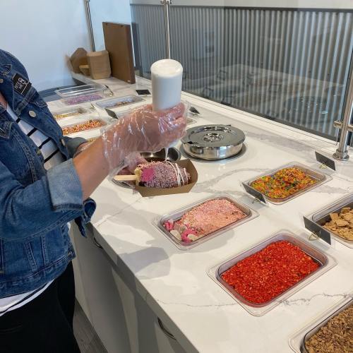 A person in a denim jacket holds a cup of white liquid over a variety of colorful toppings in containers on a counter.