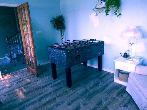 A foosball table in a light-filled room with a door, lamp, and a small table nearby.