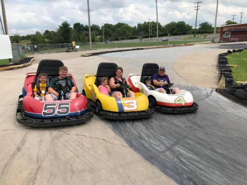 Four children and adults sit in go-karts on a racetrack, smiling and ready to race on a sunny day.
