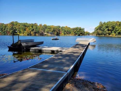 A wooden dock extends into a calm lake, with boats nearby and colorful trees reflecting in the water under a clear blue sky.