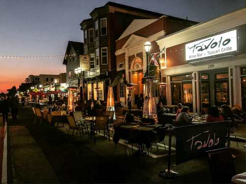 A lively street scene at dusk with outdoor dining, warm lights, and a restaurant named Tavolo.