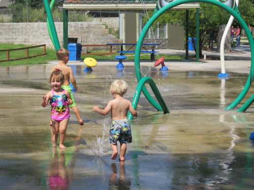 Three children play in a splash pad, running through water sprays on a sunny day.