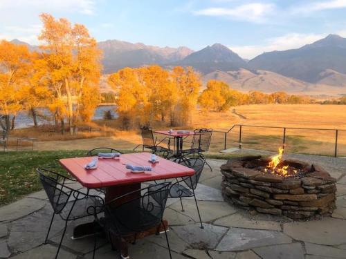 A cozy outdoor seating area with red tables, a fire pit, and autumn trees against a mountain backdrop at sunset.