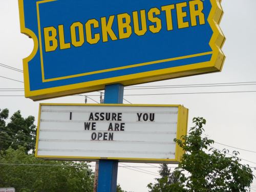 A Blockbuster sign with a message reading, "I assure you we are open," against a cloudy sky.