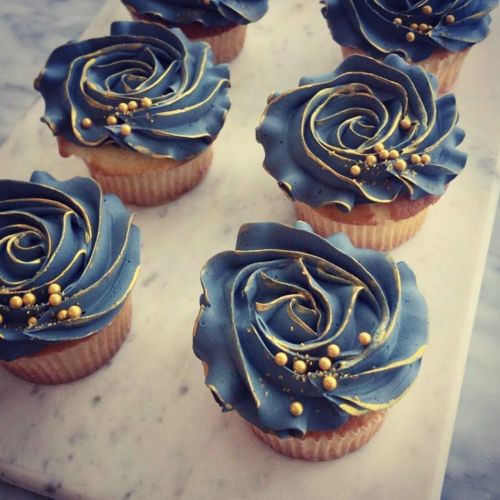 Six cupcakes with dark blue rose-shaped frosting and gold accents, arranged on a marble surface.