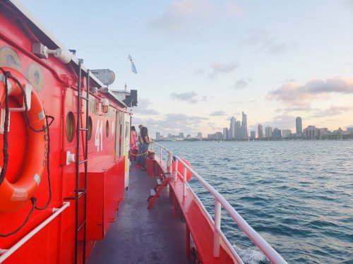 A red boat with people on deck, overlooking a city skyline at sunset with water in the foreground.