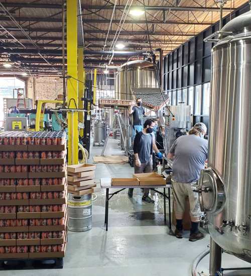 Workers in a brewery are assembling boxes and cans near large brewing equipment in a spacious industrial setting.