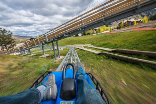 A close-up view of feet on a roller coaster track, with a blurred background of green hills and cloudy skies.