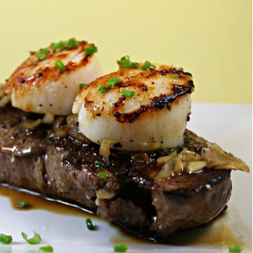 A juicy steak topped with two seared scallops and garnished with chopped green onions on a white plate.