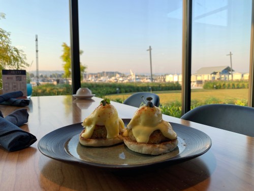 Two eggs Benedict on a plate, with a scenic view of a marina and greenery through large windows.
