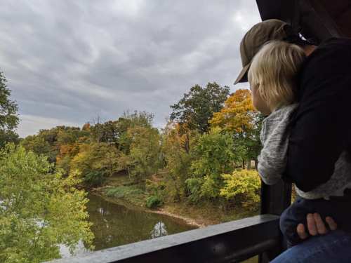 A person holds a child while looking out over a river surrounded by trees with autumn foliage under a cloudy sky.