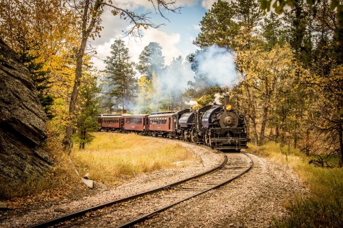 A vintage steam train curves through a forested area with autumn foliage, emitting smoke against a scenic backdrop.