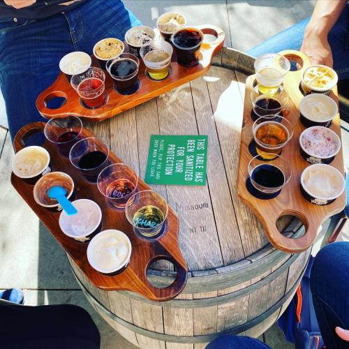 A wooden table with two flight trays of assorted beers and ice cream cups, surrounded by people’s legs.