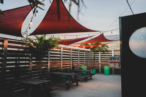 Outdoor seating area with red shade sails, wooden benches, and greenery, illuminated by string lights at dusk.