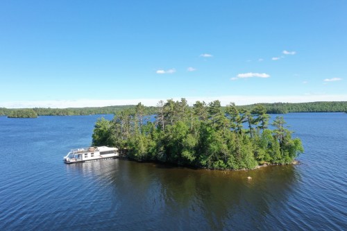 Aerial view of a small island with lush trees, surrounded by calm blue water and a houseboat docked nearby.