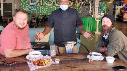 Two men sit at a table with food, while a chef stands between them, smiling in a casual dining setting.