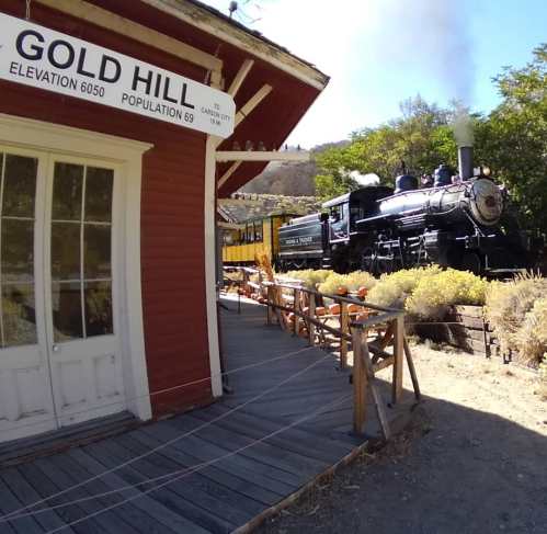 A vintage steam train near a rustic building labeled "Gold Hill," surrounded by greenery and outdoor seating.