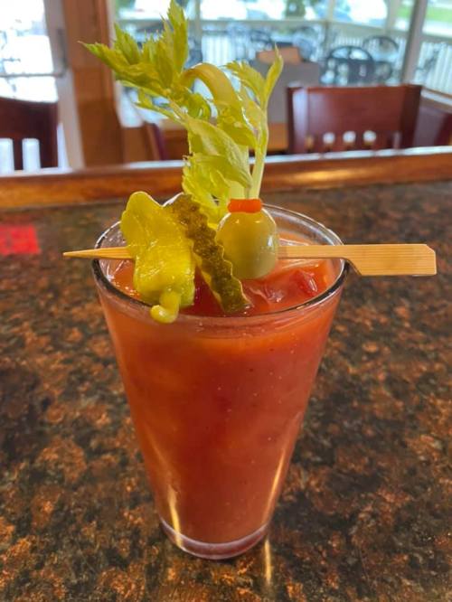 A tall glass of Bloody Mary garnished with celery, pickles, and an olive on a skewer, set on a dark countertop.