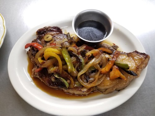 A grilled steak topped with sautéed peppers and onions, served with a small cup of sauce on a white plate.