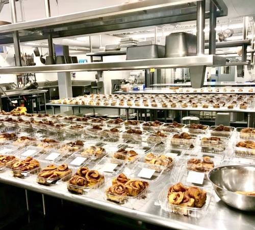 A commercial kitchen filled with trays of cinnamon rolls in clear containers, ready for distribution.