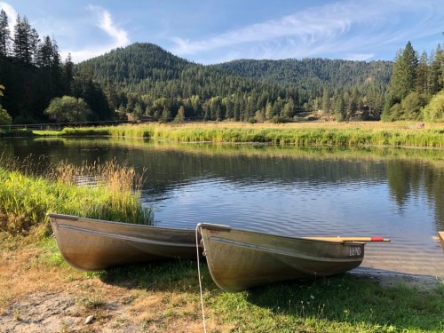 Two canoes on the shore of a calm lake, surrounded by lush green trees and mountains under a clear blue sky.