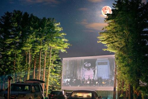 A drive-in movie screen shows a film under a full moon, surrounded by tall trees and parked cars.