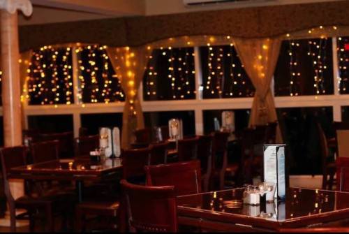 Cozy restaurant interior with wooden tables, chairs, and warm string lights illuminating the windows.