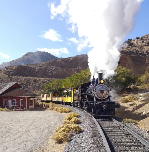 A vintage steam train chugs along a track near a rustic building, surrounded by mountains and desert landscape.