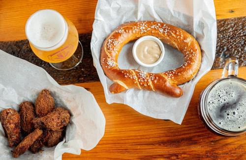 A soft pretzel with dipping sauce, chicken wings, and two glasses of beer on a wooden table.