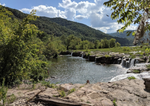 A serene landscape featuring a river, small waterfalls, and lush green hills under a partly cloudy sky.